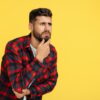 Curious bearded hipster young man in plaid shirt thinking over yellow background